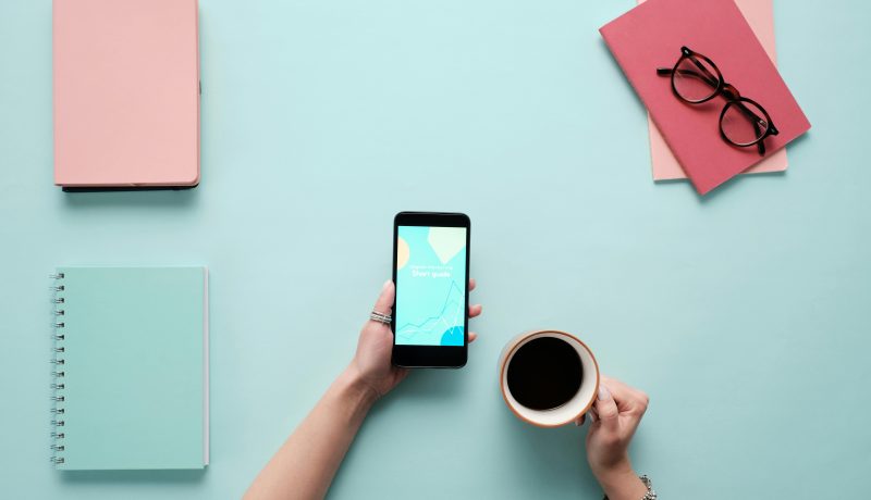 Top 5 Journaling Apps to Boost Productivity in 2025 A woman holding a phone in the left hand side and a cup of coffee on the right hand side. Books all over the table.