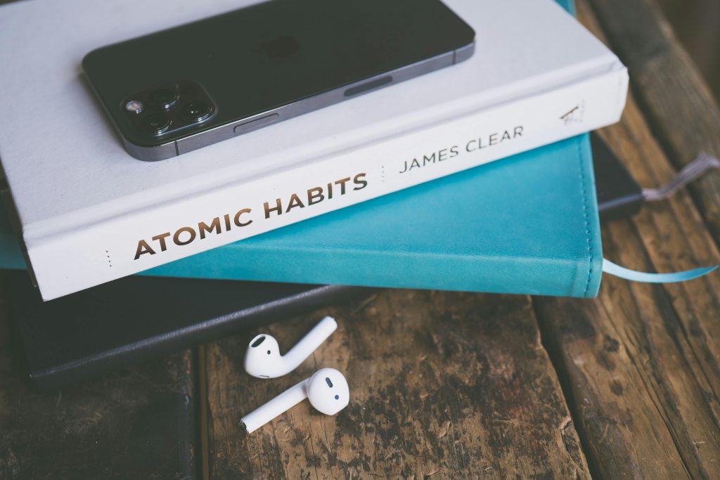 An image containing a book "Atomic Habits by Jame Clear", an iphone and airpods